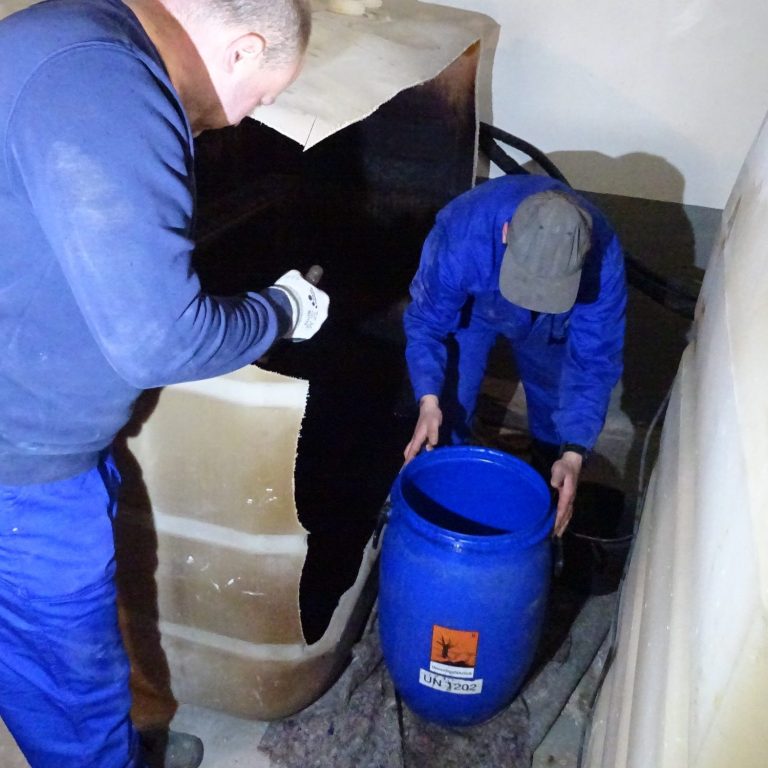 Zwei Männer in blauen Overalls entleeren einen zerschnittenen Heizöltank in ein blaues Fass.