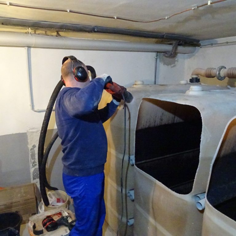 Ein Mann in einem Blauen Pulli und Latzhose demontiert einen alten Heizöltank im Keller